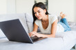 © F8  \ Suport Ukraine - Young smiling woman lying down the bed in front of her laptop with her legs raised slightly