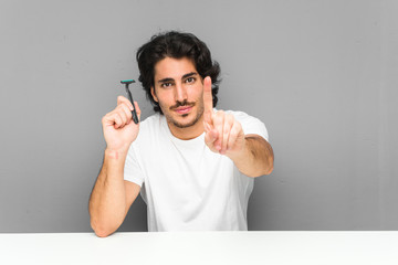 Wall Mural - Young man holding a razor blade showing number one with finger.