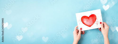 Female hands holding card with a painted red heart on blue background with bo...