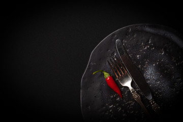 Closeup round ceramic plate with pattern of circles, vintage fork, red fresh chili pepper on black background. Concept modern shooting of advert menu from chef for restaurant, tableware catalog