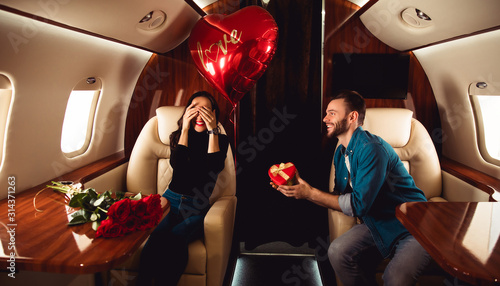 Love in the air. A beautiful couple is celebrating St Valentine’s Day on a private jet. A man is giving a present in a red box to his girlfriend, who is sitting with her eyes closed.