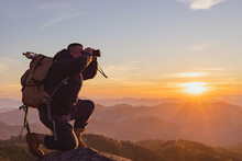 Young Man With Backpack And Holding A Binoculars Sitting On Top Of Mountain At Sunset