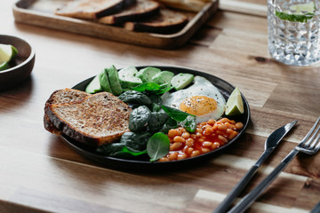 Healthy breakfast or lunch at home or cafe with fried egg, avocado, toasts, beans and fresh spinach on a wooden table.