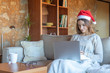 © Marx Bamala - Serious young woman with Santa hat watching a movie sitting on the sofa at home in front of a laptop