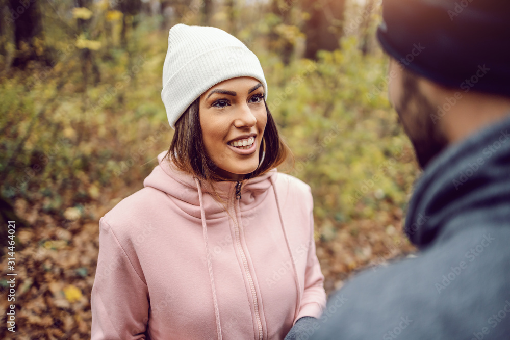 Pretty smiling young woman in sportswear, with beanie on head standing in nature,taking a break and talking to her friend. Fitness in nature on cold weather concept. - obrazy, fototapety, plakaty 