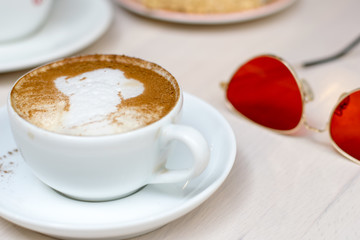 hold ceramic white with cappuccino above wooden plate and rustic table. Milk foam on top in heart shape