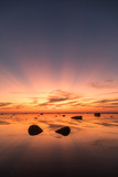 Fototapeta Desenie - Seascape view with the shallow calm seawater with erratic boulders and sunset colored sky with afterglow sun rays in the sky and reflecting on water