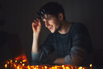 Canvas Print - Portrait of young man wearing glasses, looking surprised in laptop, dark room with garlands at home.