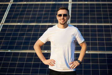 Wall Mural - Portrait of a young successful man standing smiling with his hands on waist, wearing white t-shirt and dark sunglasses, in front of a big solar pv module