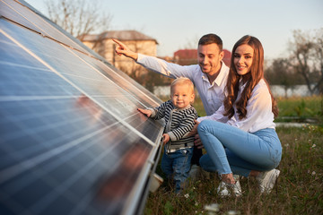 a young family of three is crouching near a photovoltaic solar panel, smiling and looking at the cam