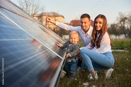 Fotografía A young family of three is crouching near a photovoltaic solar panel, smiling an