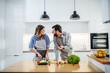 © Dusan Petkovic - Young charming smiling caucasian woman in apron standing in kitchen and cutting cucumber while talking with her boyfriend. Man holding cherry tomato and talking about healthy lifestyle.
