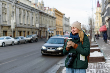 Girl in winter clothes with camera and map in the historical center of the city is holding a cup of coffee on the street