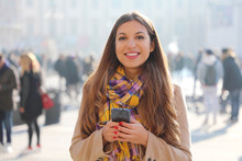 Happy Beautiful Young Woman Looking At Camera And Holding Mobile Phone Outdoor With Blurred Crowd Of People On Street Background, Selective Focus. City Lifestyle People Technology.