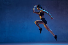 Caucasian Young Female Athlete Practicing On Blue Studio Background In Neon Light. Close Up Of Sportive Model Jumping High, Running. Body Building, Healthy Lifestyle, Beauty And Action Concept.