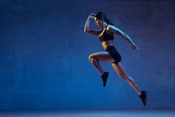 Caucasian young female athlete practicing on blue studio background in neon light. Close up of sportive model jumping high, running. Body building, healthy lifestyle, beauty and action concept.