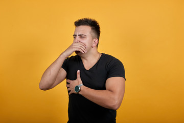 Sickness young man wearing black shirt isolated on orange background in studio covered mouth with hand, cough, keeping hand on chest. People sincere emotions, lifestyle concept.