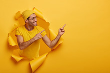 Positive European Male Model Points Right With Both Index Fingers, Suggets Try To Use Product, Turns Aside, Stands In Ripped Paper Hole, Wears Yellow Hat And T Shirt. Check Out Amusing Promotion