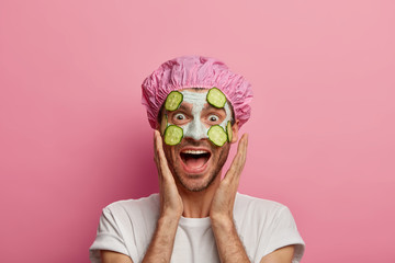 Photo of funny male model touches cheeks, laughs happily, enjoys freshness of skin, has regular beauty treatments, wears facial mask with cucumber slices, wears shower cap and t shirt, stands indoor