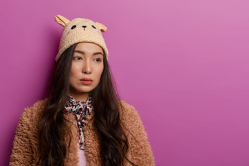 Wall Mural - Sad brunette woman turns from camera, has seious thoughtful expression, wears hat and coat, feels lonely and discontent, stands against lilac wall, copy space for your promotion. Pensive Asian girl