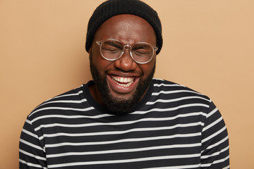 Wall Mural - Headshot of dark skinned bearded man laughs out happily, squints from joy, shows white teeth, wears black hat and striped sweater, isolated over brown background, hears hilarious joke, has fun