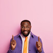 © WHstudio Leushin N - Cheerful unshaven man points both index fingers up, demonstrates successful idea, promots copy space, wears spectacles, orange tie and purple coat, isolated over rosy background, introduces promo