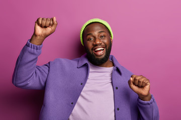 Wall Mural - Overjoyed dark skinned man clenches fists while dances, being on disco party, enjoys funny music, has fun, laughs with broad smile, wears stylish bright outfit, poses at studio. Good emotions concept