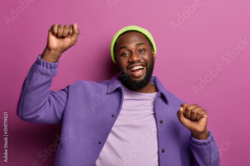 Overjoyed dark skinned man clenches fists while dances, being on disco party, enjoys funny music, has fun, laughs with broad smile, wears stylish bright outfit, poses at studio. Good emotions concept