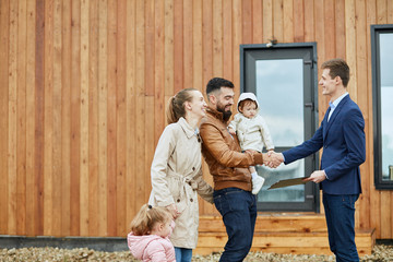 Caucasian happy family of four and realtor wear blue suit stand outdoor near new house just sold. Men shake hands.