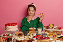 Oops, So Much Calories. Surprised Korean Woman Holds Tasty Pastry, Eats Cheat Meal Containing Much Sugar, Has Imbalanced Nutrition And Diet Breakdown, Poses Near Table With Various Desserts.