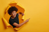 Fototapeta  - Pleased dark skinned Afro American woman stands in ripped background, laughs happily, poses in paper hole, points on right side, isolated on yellow wall. Space for your advertising or promotion.