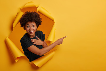 happy curly haired african american woman laughs positively, points aside on copy space, wears black