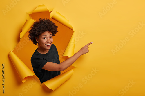 Fotomural African American woman in black t shirt, stands in paper hole, points away on blank space, stands in ripped paper, has cheerful expression, isolated over yellow background