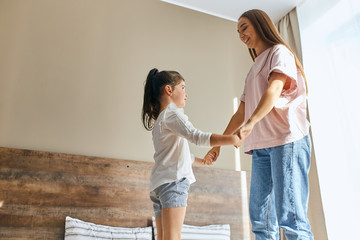 Two beautiful sisters dressed in casual outfit, standing on bed in brightly lighted bedroom, holding hands, looking at each others with happy face, ready to jump, shot from below, family concept