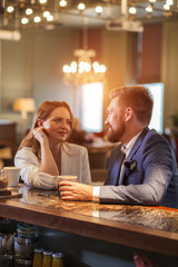 Close up of couple having first date in romantic place. Beautiful young woman looking at boy friend with admire, lovely man holds a cup of coffee, turns head towards pretty girl with kind smile