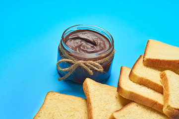 Single glass transparent jar with rope full of melted chocolate or cream near scattered pieces of vanilla bread lies on blue desk on kitchen. Concept of nutritious breakfast or lunch