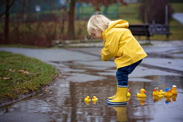 Beautiful funny blonde toddler boy with rubber ducks and colorful umbrella, jumping in puddles and playing in the rain