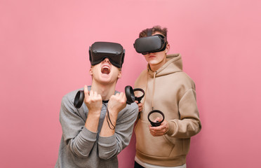 Portrait of emotional 2 young people in VR helmets on a pink background playing virtual reality games on skaters in hands. Couple of expressive friends playing VR games on console.