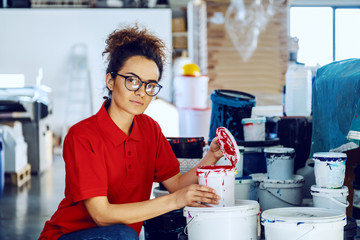 Beautiful caucasian female employee crouching next to bucket with colors and opening red color. Printing shop interior.