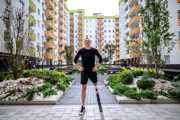 Wall Mural - Full length of handsome smiling sportsman with artificial leg standing with hands on hips outdoors surrounded by buildings. Back light.