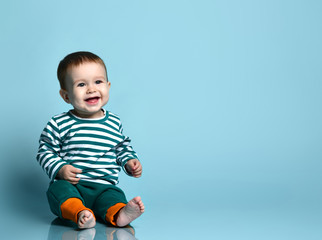 Little baby boy in stylish casual clothing barefoot sitting on floor and smiling over blue wall background