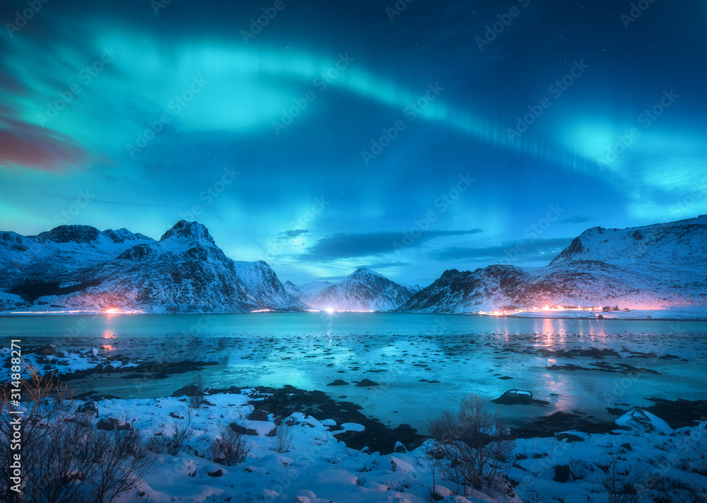 Aurora borealis over the sea coast, snowy mountains and city lights at night. Northern lights in Lofoten islands, Norway. Starry sky with polar lights. Winter landscape with aurora reflected in water - obrazy, fototapety, plakaty 