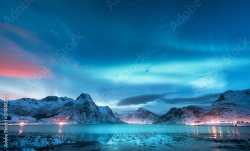 Fototapeta Zorza polarna nad morzem, zaśnieżone góry i światła miasta nocą. Zorza polarna na Lofotach, Norwegia. Gwiaździste niebo z polarnymi światłami. Zimowy krajobraz z zorzą odbijającą się w wodzie