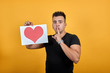 Caucasian young man wearing black shirt isolated on orange background in studio keeping picture with red heart, doing shh gesture with his finder. People sincere emotions, lifestyle concept.