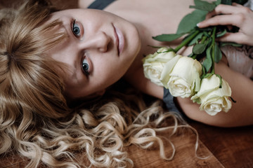 portrait of a beautiful young adult girl with blue eyes and blond curly hair lying with white roses