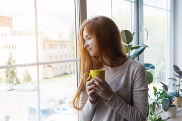 candid portrait of young smiling red haired woman holding yellow coffee cup, enjoying morning coffee and new day