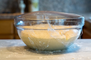 A Clear Glass Bowl of Rising Bread Dough Covered with Plastic Wrap on a Kitchen Counter 