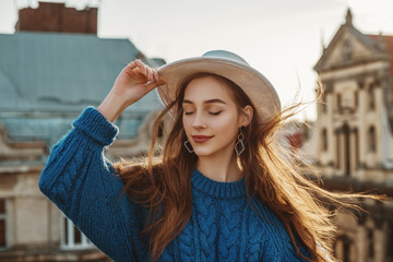 Outdoor close up fashion portrait of young happy brunette woman with long natural hair. Model wearing blue sweater, stylish white hat, trendy earrings, posing at sunset, in European city. Copy space