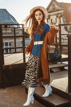 Fashionable  Woman Wearing Trendy Brown Faux Fur Coat, Blue Knitted Sweater, Wide Belt, Stylish  Hat, Pleated Zebra Print Skirt, White High Boots, Holding Hobo Bag, Posing At Sunset, In European City