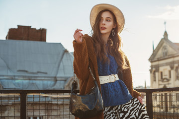 Fashionable  woman wearing trendy brown faux fur coat, blue knitted sweater, wide leather belt, stylish white hat, holding hobo handbag, posing at sunset, in European city. Copy, empty space for text